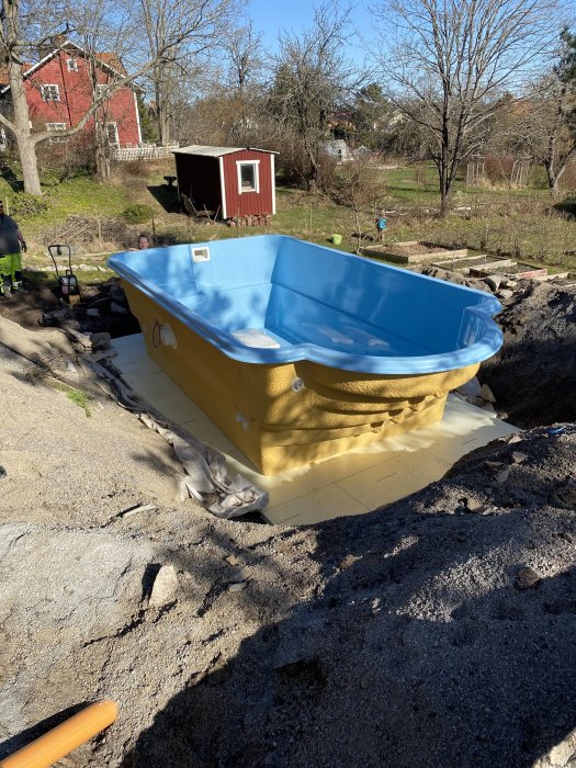 En gul och blå pool placerad på en presenning i en grävd grop i en trädgård, omgiven av grus och byggmaterial.