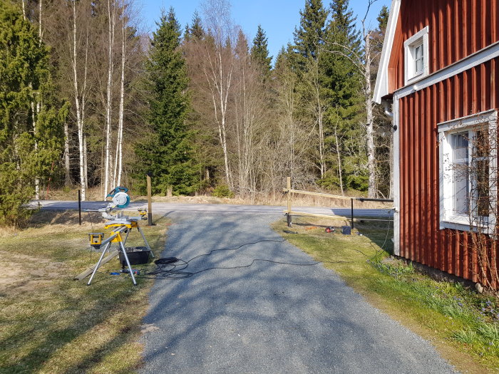 Uppställda staketreglar i våg vid en röd stuga med en kap- och gersåg i förgrunden och ett skogsbryn i bakgrunden.