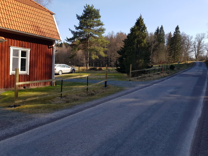 Nyinstallerade staketreglar längs en grusväg med röd stuga och bil i bakgrunden och skog runtomkring.