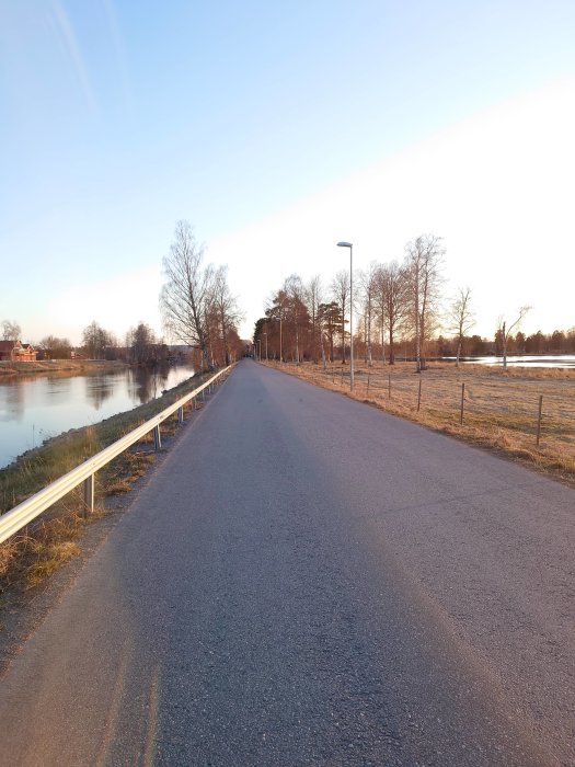 Asfalterad gång- och cykelväg längs med en flod omgiven av träd och gräs i skymningsljus.