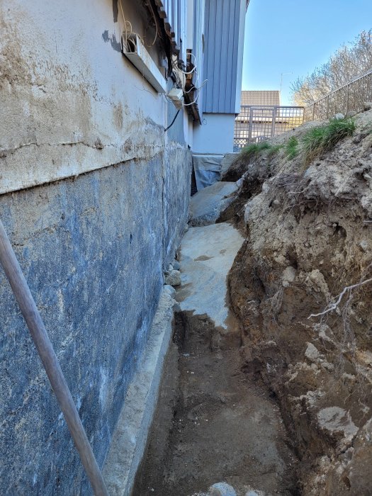 Grävd grund längs husvägg för dränering och isolering, visar berg och jord nära bostadens fundament.