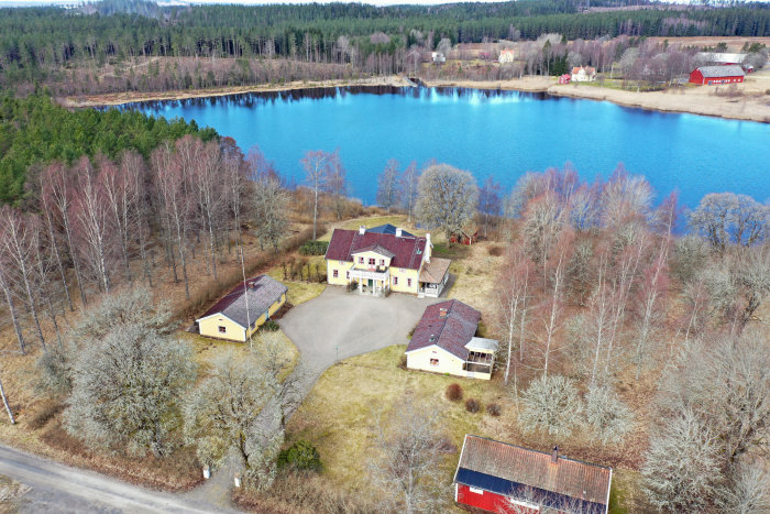 Flygbild över en herrgårdsliknande fastighet med outbyggnader intill en blå sjö omgiven av träd.