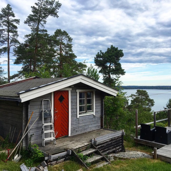 Nybyggd bastu med relaxavdelning, röd dörr och utsikt över vatten, omgiven av träd och natur.