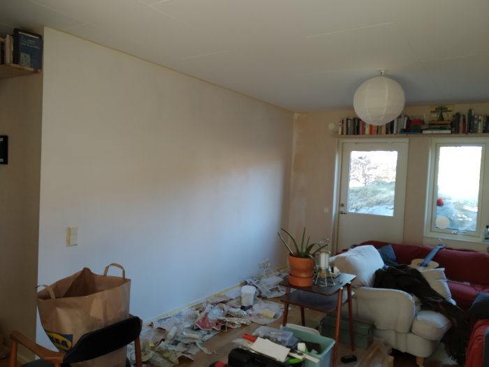 Vägg med ojämn vitmålning och halo-effekt i solljus, i ett rörigt vardagsrum under renovering.
