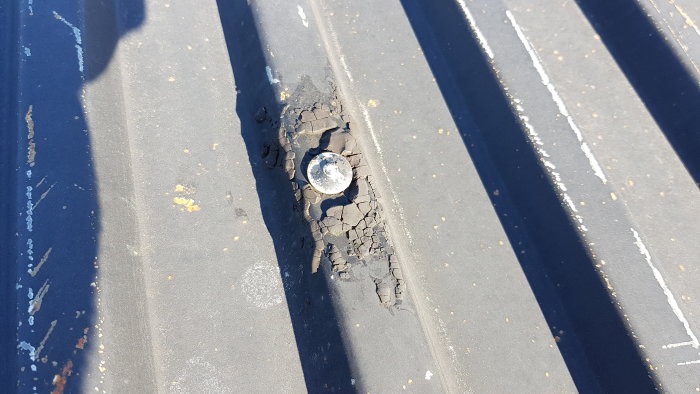 En spik omgiven av sprucken svart massa på ett plåttak.