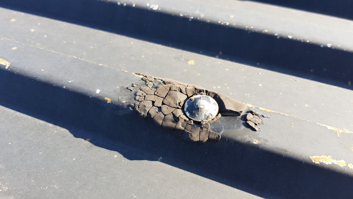 Svart, sprucken massa runt en spik på ett plåttak, möjligen gammal tätning eller tjära.