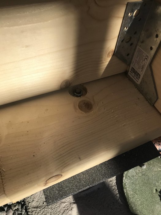 Detaljbild på en träkonstruktion med två grova bjälkar och en metallhörnplåt i solljus som en del av en byggstomme.