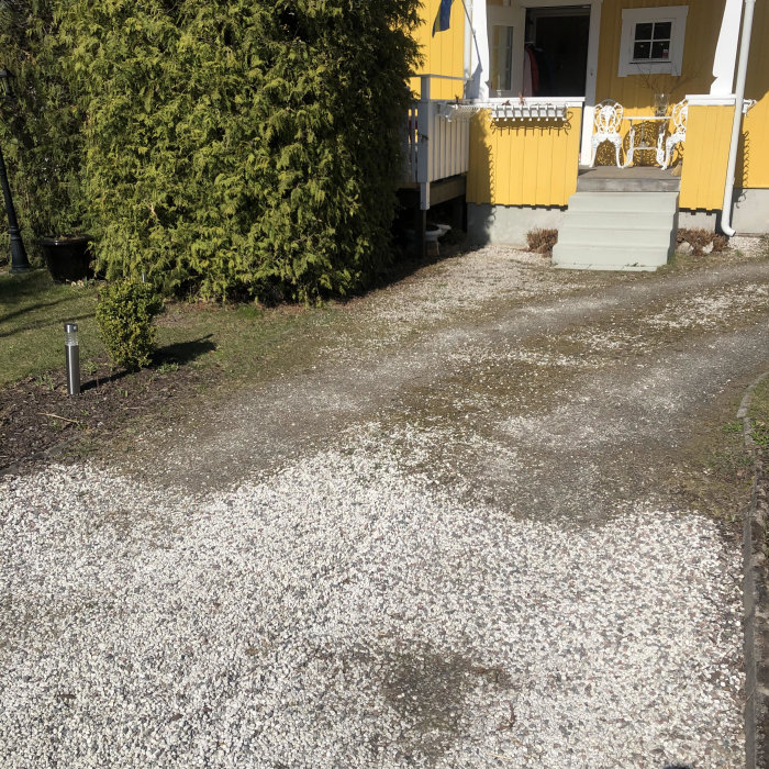 Grusgång med inslag av dolomit som behöver underhåll framför ett gult hus med gröna buskar.