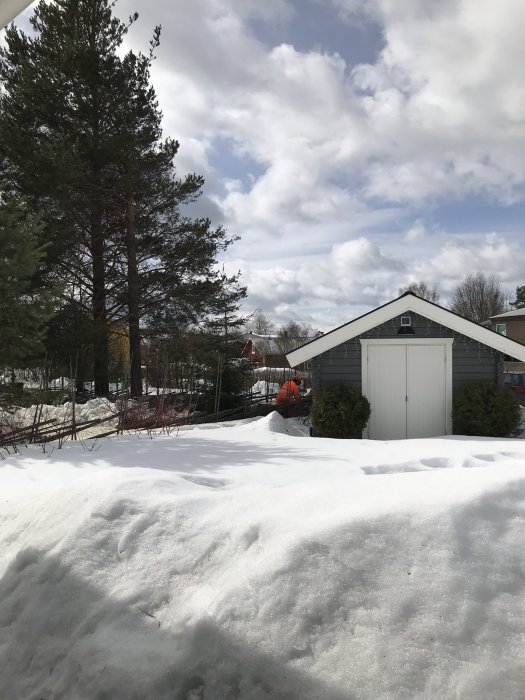 Vit snö framför en grå garagebyggnad med träd och molnig himmel i bakgrunden.