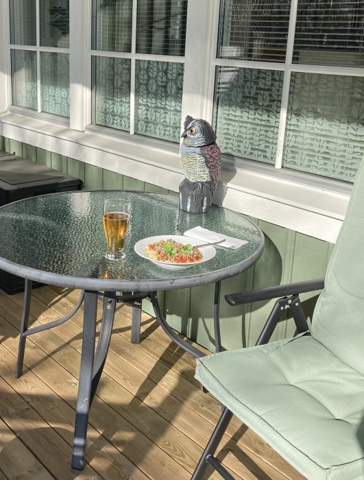 Ugglefigur sitter vid ett utebord med ett glas öl och en tallrik mat på en solig veranda, med stolar och fönster i bakgrunden.