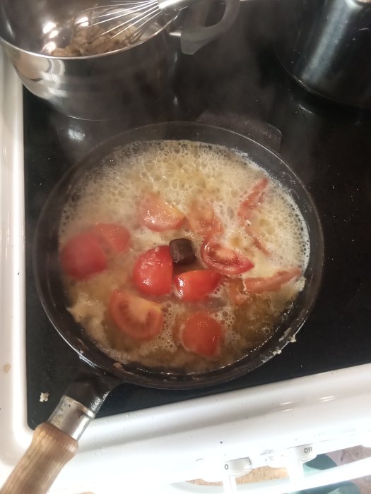 Stekpanna på spis med buljong, tomatbitar och lök under tillagning av hembakad sås.