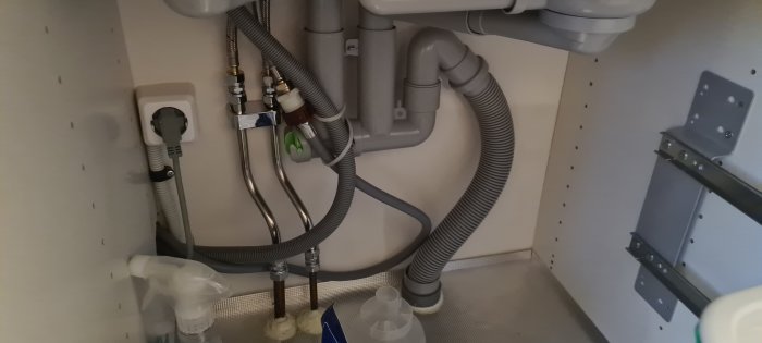 Undersida av köksbänk med synliga avloppsrör, slangar, eluttag och sopbehållarskena.