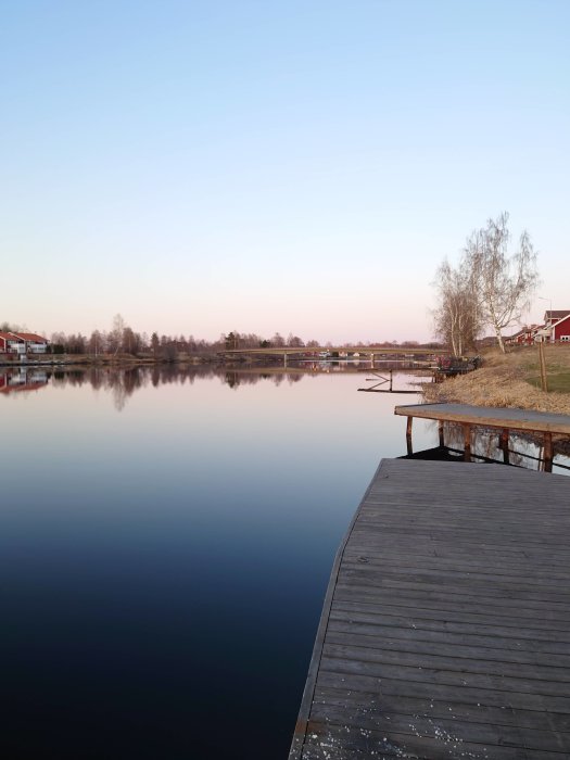 Träbrygga som leder ut i en spegelblank sjö vid skymning med träd och hus i bakgrunden.