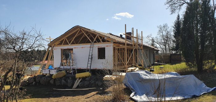 Ett hus under konstruktion med vindpapp på väggarna, byggmaterial på marken och en tydlig blå himmel.