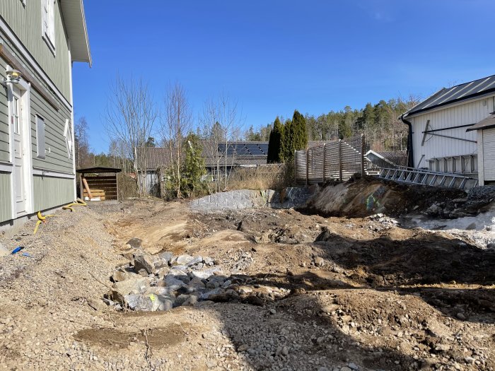 Ojämn och stenig byggplats med lutande berggrund nära husvägg, liten makadam och grävmaskiner.