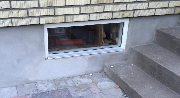 Ett källarfönster med en aluminiumlist längst ner för att förhindra vattenintrång, intill en trappa.