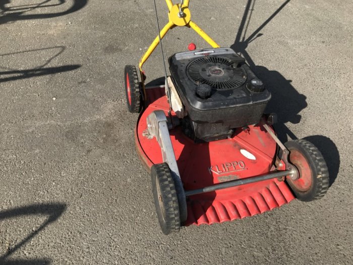 Röd Klippo gräsklippare med gul handtag syns på asfalterad yta.