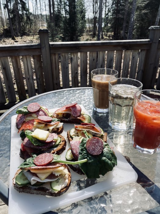 Mackor med salami, ost och grönsaker på en tallrik, med ett glas kaffe och vatten på ett utebord i solljus.