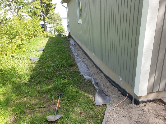 Pågående markarbete vid hus, med grävd kant, fiberduk, grus och spade på gräsmattan.