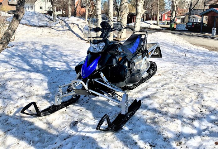 Blå och svart snöskoter parkerad på snötäckt mark med träd och hus i bakgrunden.