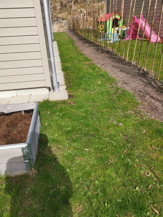 Trädgårdssida med pallkrage vid husvägg, gräsmatta, staket och lekstuga i bakgrunden.