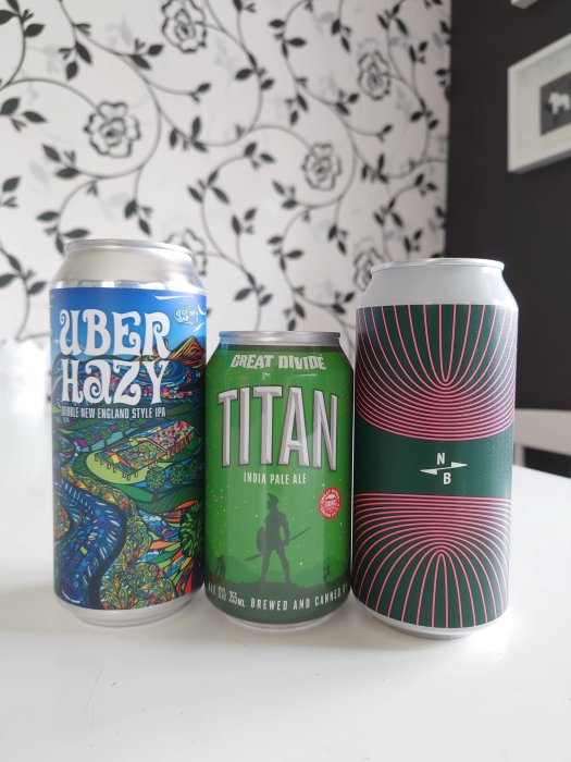 Tre ölburkar på en vit yta framför tapet med blomstermotiv, två IPAs, en märkt "Uber Hazy", den andra "Titan".