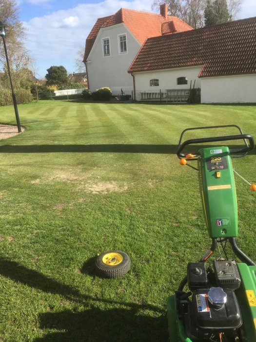 Grön gräsklippare i förgrunden med nyslagen gräsmatta och hus i bakgrunden.