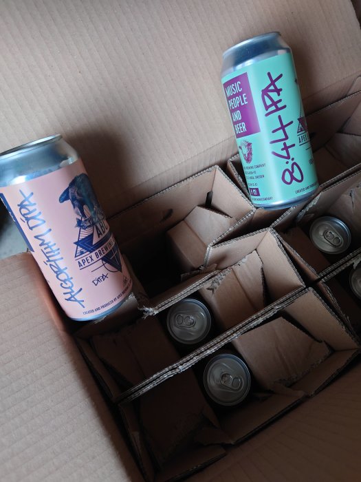 Öppen wellpapplåda med flera ölburkar från Apex Brewing, två synliga med färgglada etiketter.