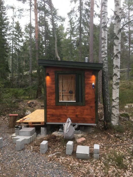 Påbörjat bygge av en liten stuga med träfasad i skogsmiljö, står på betongblock, med tillfällig elanslutning och byggmaterial framför.