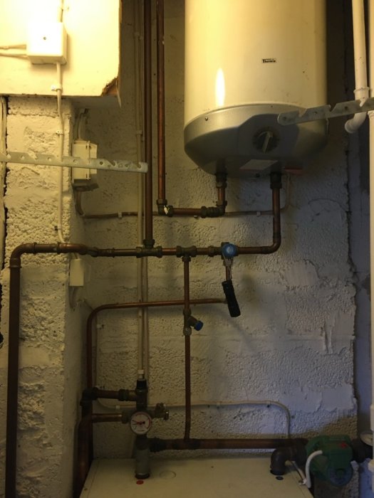 Ett litet pannrum med äldre varmvattenberedare, kopparledningar och ventiler på en murad vägg.