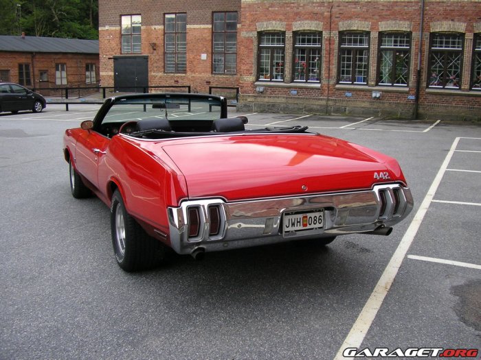 Röd Oldsmobile 442 Convertible parkerad på en gata med byggnader i bakgrunden.