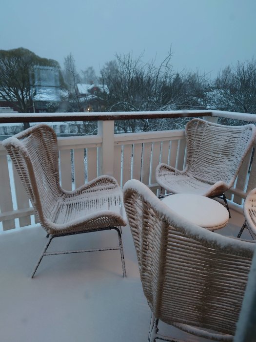 Två rottingstolar täckta med snö på ett balkongdäck med ett dimmigt vinterlandskap i bakgrunden.