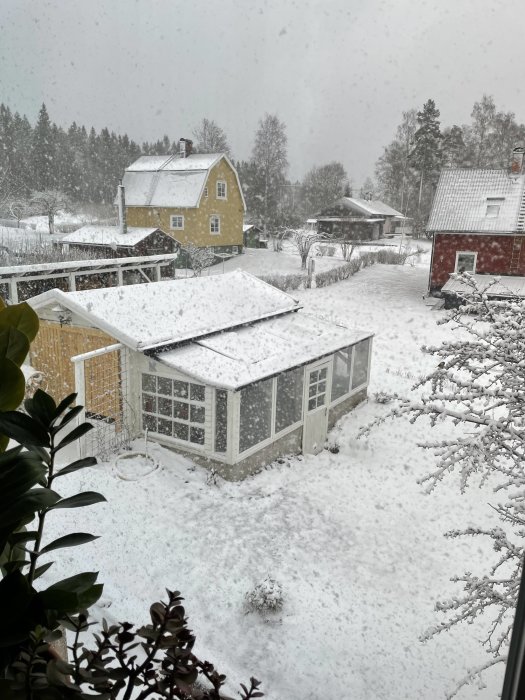 Uteplats med inglasning under snöfall, omgiven av snötäckt trädgård och hus i bakgrunden.