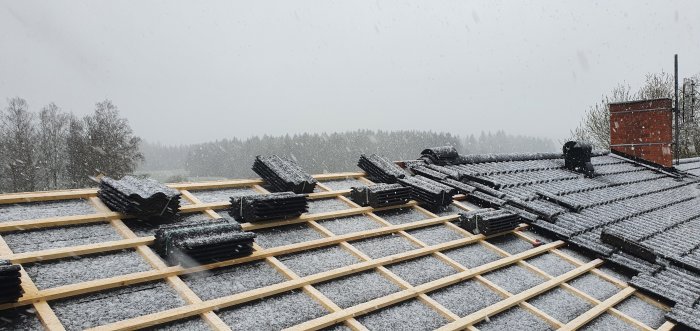 Snötäckt byggarbetsplats med takstolar och några staplar av takpannor, snöfall i bakgrunden.