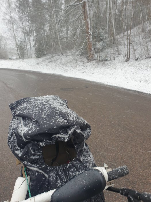 Skadad svart barnvagn täckt med snö på en vägkant vid ett snöigt skogslandskap.