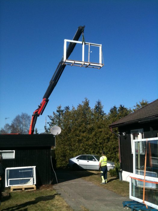 Lyftkran höjer ett stort fönsterparti ovanför hus på klarblå himmel.