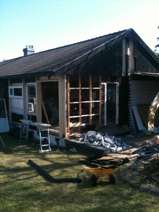 Hus under renovering med öppen vägg och byggmaterial utspridda på gräsmattan.