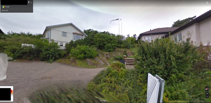 Vy över en gata med ett vitt hus till vänster och ett annat hus med en svensk flagga på höger sida, grönska i förgrunden.