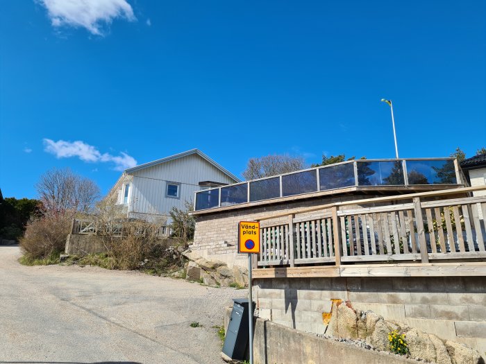 Trädäck med glasräcke vid hus, gata och 'Vändplats' skylt, under en klarblå himmel.