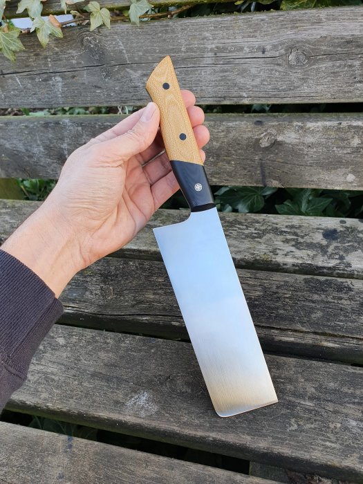 Hand håller en nyligen färdigställd högre nakiri-kniv med konvex geometri mot en träbänk.