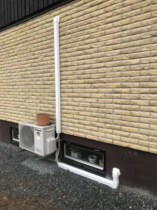 Vägg med luftvärmepump och ett ventilationsrör som går upp längs tegelfasaden till ett hus.
