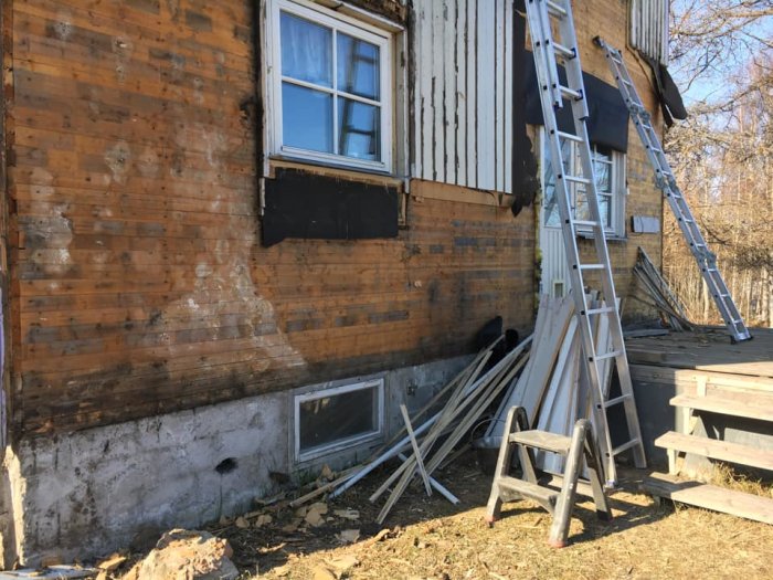 Delvis avtäckt husvägg med synliga mögelskador på spontad undervägg och byggmaterial inklusive stege framför.