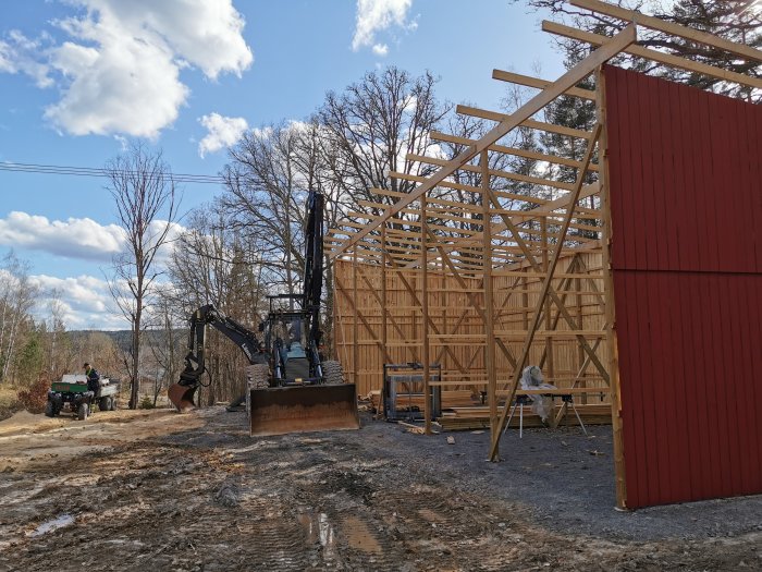 Byggplats med grävmaskin och vedbod under konstruktion, delvis rödmålad vägg i soligt väder.