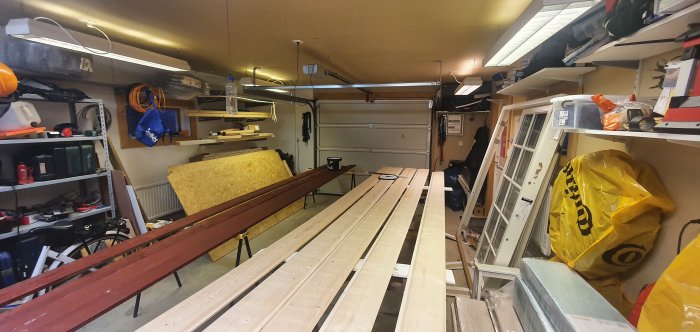 Oorganiserat garage med staplar av omålade panelbrädor redo för målning och diverse byggmaterial och verktyg runtomkring.