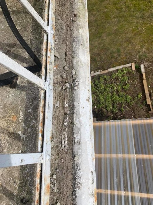 Balkongvy med slitet räcke och skadad betong, i behov av renovering, med trädgård nedanför.