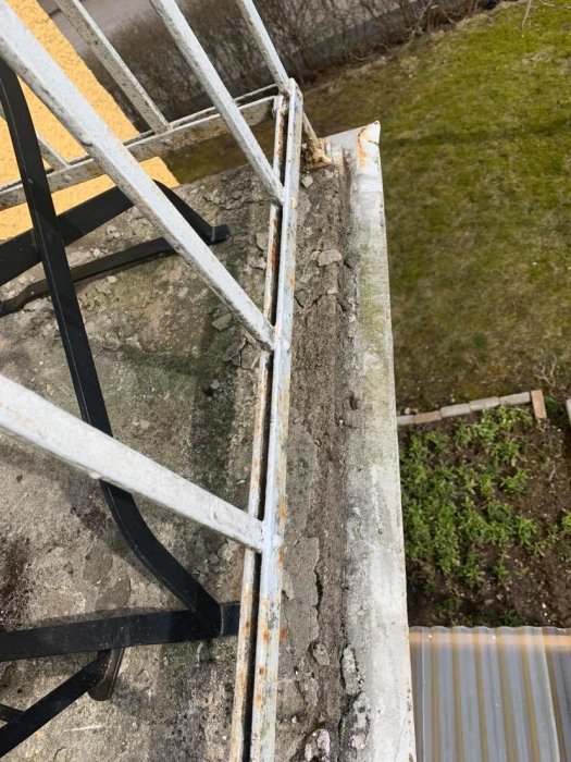Sliten balkongkant med delvis trasigt betong och rostigt, vitmålat räcke, behov av renovering syns.