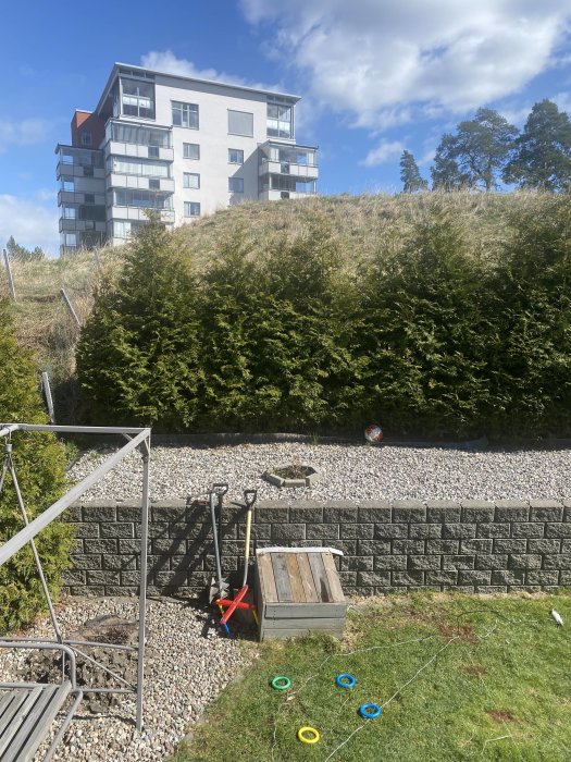 Trädgård med stödmur och gräsmatta framför flervåningshus, markering vid plinthål nära mur och leksaker.