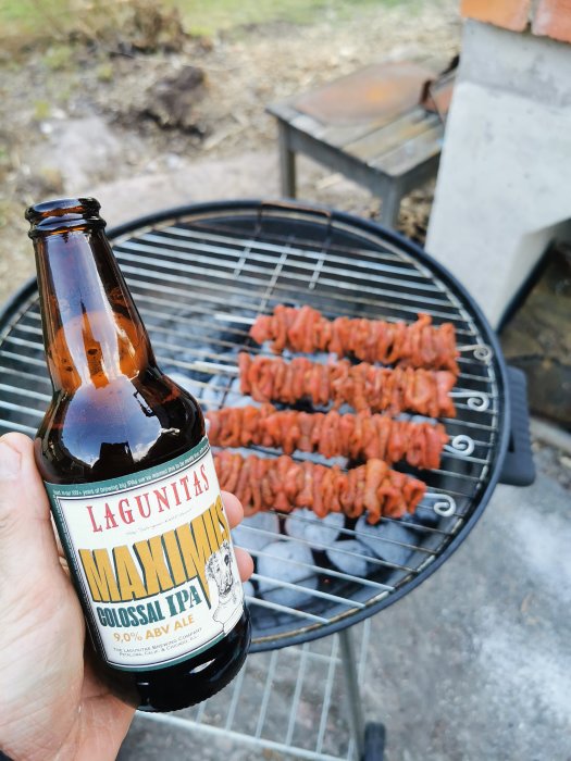 Hand håller en flaska öl framför en grill med köttspett.