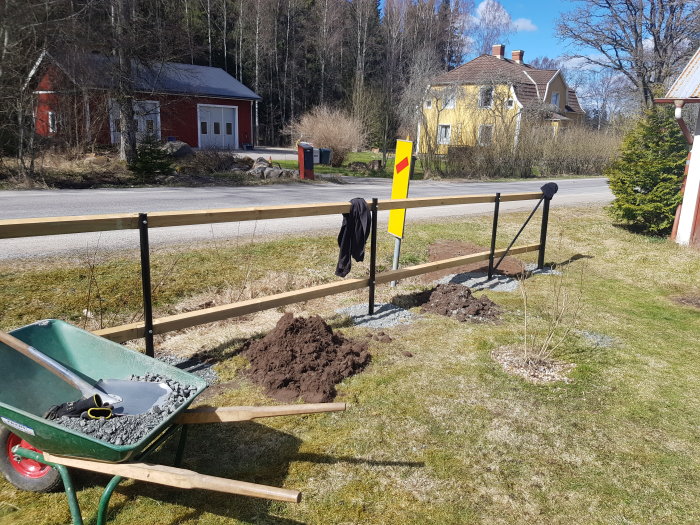 Staketbygge med nygrävda stolparhål fyllda med grus bredvid en tom grusgång och skottkärra med verktyg.