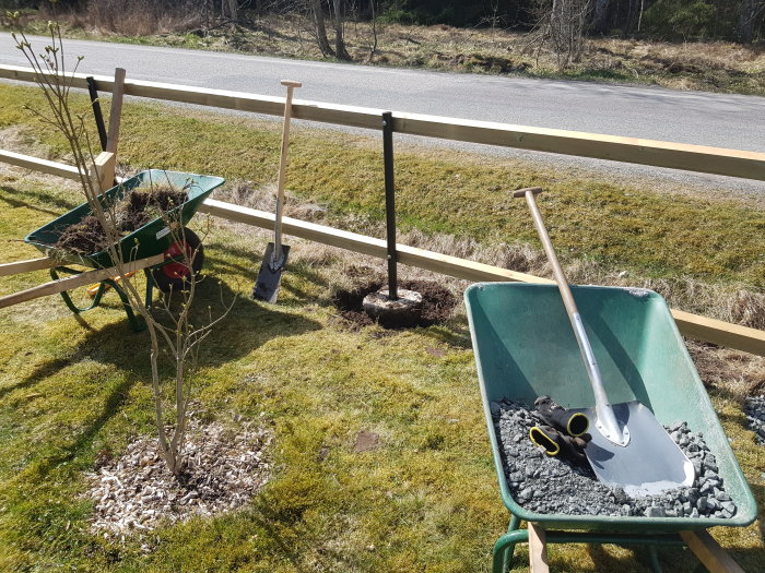 Trädgårdsvagn fylld med jord, en spade och en kratta bredvid ett hål, samt en vagn med grus och en skyffel, i arbetet med att förstärka staketstolpar.